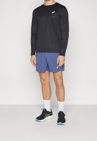 Mustat pitkät hiha-atletikkapaita, jossa on valkoinen logo, yhdistettyinä sinisiin shortseihin. Päällä mustat ja vihreät urheilukengät ja valkoiset sukat.