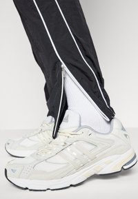 Zwarte trainingsbroek met witte zijkanten, voorzien van ritsen bij de enkels, gecombineerd met lichtgrijze sportschoenen met mesh- en suède details.