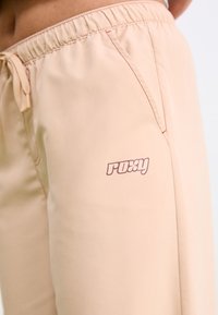 Light peach sweatpants with an elastic waistband, side pockets, and a small purple Roxy logo on the left thigh. Soft fabric, smooth texture.