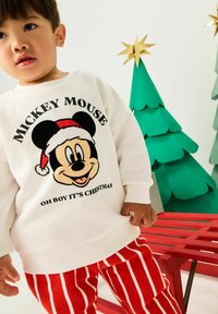 Fehér pulóver fekete "Mickey Mouse" grafikával és piros Mikulás sapkával; piros-fehér csíkos bolyhos nadrággal párosítva; zöld papír karácsonyfák.