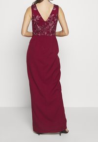 Burgundy aftonklänning med en strukturerad, broderad bodice och en V-formad ryggdesign. Lång, flödande kjol med en slät finish, bärs med klackar.