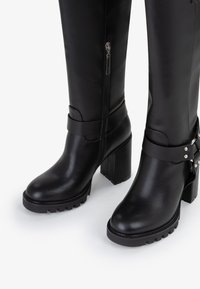 Stivali neri al ginocchio in pelle liscia, con tacco a blocco, zip laterale e una cintura regolabile con hardware argentato.