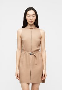 Γυναίκα που φοράει μπεζ φόρεμα χωρίς μανίκια με λεπτή μαύρη κάθετη γραμμή και ταιριαστή ζώνη με ασημί αγκράφα, στέκεται μπροστά σε λευκό φόντο.