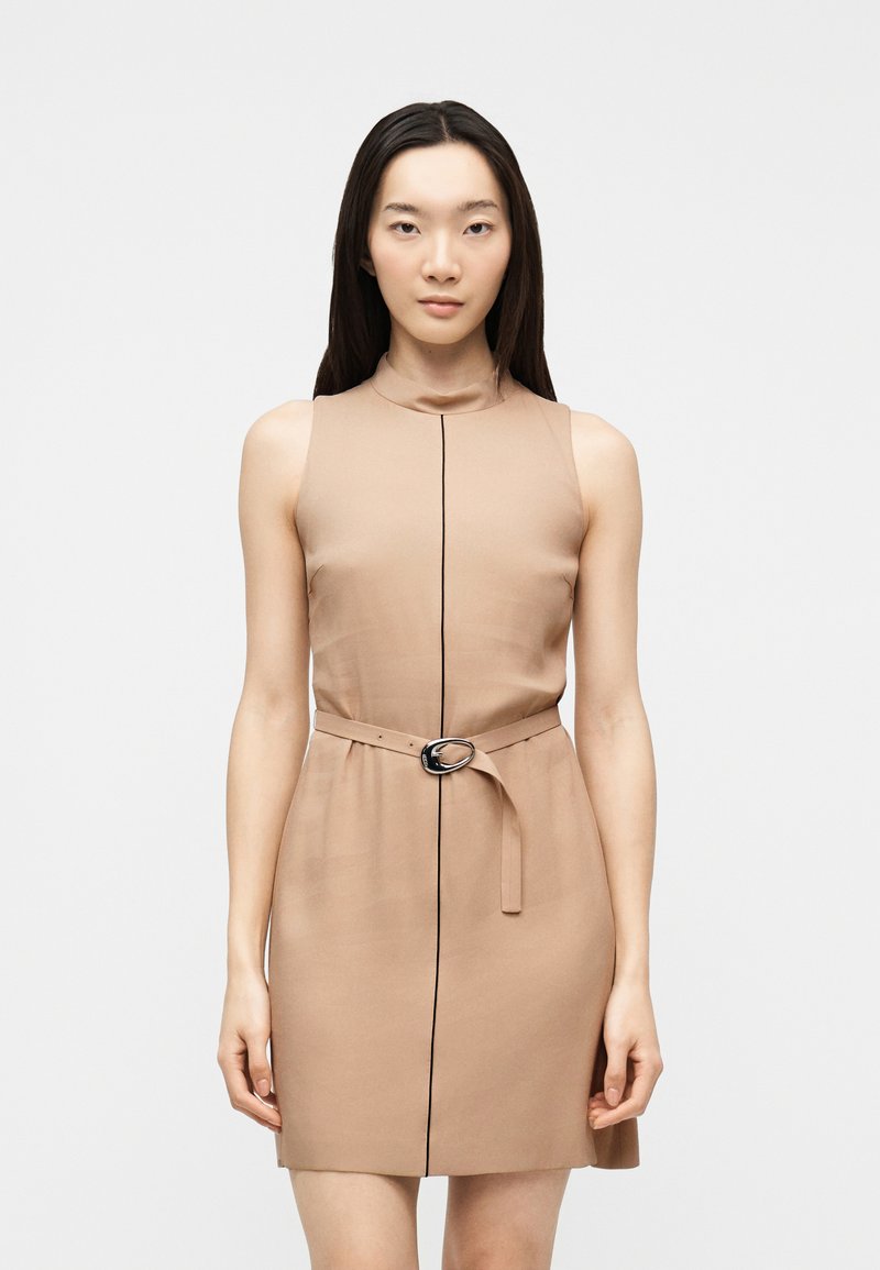 Γυναίκα που φοράει μπεζ φόρεμα χωρίς μανίκια με λεπτή μαύρη κάθετη γραμμή και ταιριαστή ζώνη με ασημί αγκράφα, στέκεται μπροστά σε λευκό φόντο.