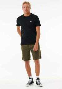 Zwarte katoenen t-shirt met een klein logo, gecombineerd met olijfgroene corduroy shorts en zwarte high-top sneakers. Effen achtergrond.