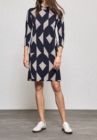 Robe bleue marine et grise à motifs géométriques, manches 3/4, longueur genou. Associée à des baskets blanches sur un fond de sol gris.