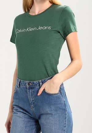 Grünes Baumwoll-T-Shirt mit Rundhalsausschnitt und weißem "Calvin Klein Jeans" Schriftzug. Kombiniert mit hochgeschnittenen blauen Jeans aus Denim.