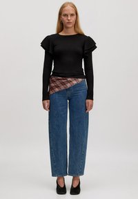 Black ribbed long-sleeve top with ruffled shoulders paired with wide-leg blue jeans; layered red and gray plaid wrap detail at waist.