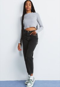 Pelēks garo piedurkņu apgriezts tops, melnas augstās jostasvietas džinsus un kedas ar zaļiem akcentiem. Braidots mati un stāvoša poza uz gaiša fona.