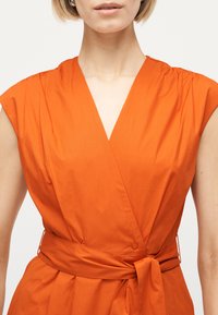 Γυναίκα που φοράει αμάνικο πορτοκαλί φόρεμα τύπου wrap με V-λαιμόκοψη και ζώνη στη μέση, εμφανίζοντας τον λαιμό και τον άνω κορμό.