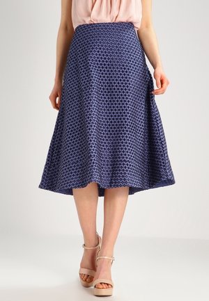 Mujer con falda midi azul marino estampada y sandalias de tacón beige, de pie contra un fondo claro y liso.