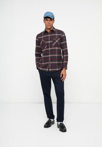 Geruite overhem dress shirt in rood en marineblauw, twee voorkeurszakken, gecombineerd met marineblauwe broek en zwarte instappers. Lichte blauwe pet maakt de outfit compleet.