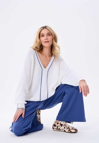 Witte gebreide trui met een V-hals en blauwe strepen, gecombineerd met losse blauwe broeken en bruin-witte sneakers met een patroon.