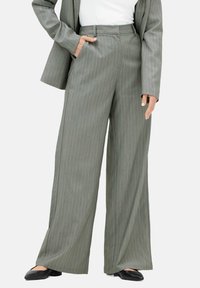 Kvinne iført grå stribede vide bukser, matchende blazer, hvid top og sorte spidse flader, med den ene hånd i lommen.