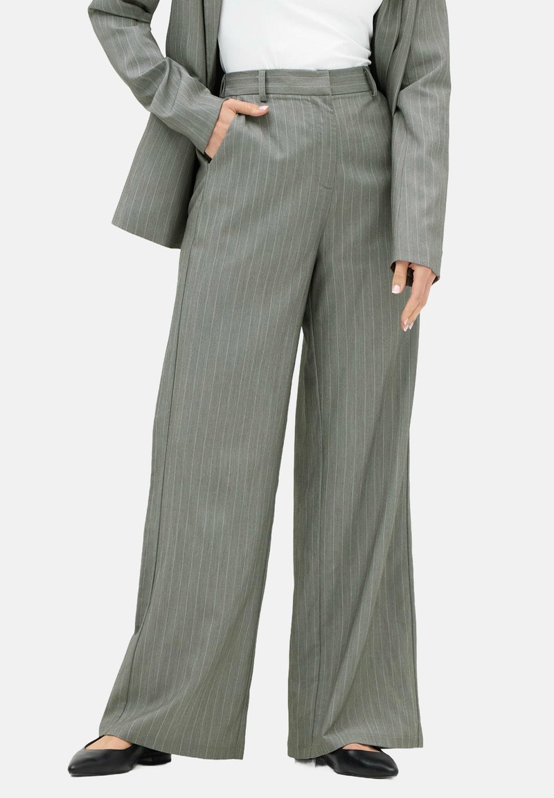 Kvinne iført grå stribede vide bukser, matchende blazer, hvid top og sorte spidse flader, med den ene hånd i lommen.