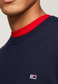 Donkerblauwe gebreide trui met een rode kraag; voorzien van een klein geborduurd logo op de borst; gestructureerde stof en ronde halslijn.