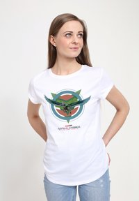 Camiseta de algodón blanca con un gráfico colorido de un superhéroe volando contra un emblema circular, con acentos verdes y naranjas.