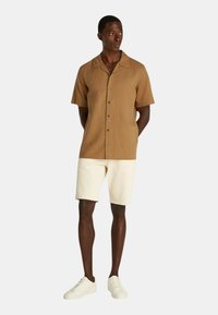 Camicetta marrone a maniche corte con colletto, abbinata a pantaloni corti beige chiaro e sneakers bianche. Tessuto liscio e vestibilità rilassata.