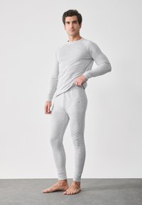 Man som bär en ljusgrå åtsittande termisk långärmad topp och leggings, står barfota på grått golv mot en vit vägg.