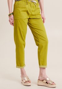 Groene katoenen broek met een elastische tailleband, twee zijzakken en opgerolde mouwen. Gecombineerd met gouden sliders op een gestructureerd plateau.