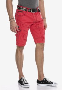 Rote Cargo-Shorts mit mehreren Taschen, genähten Details und einem schwarzen Nieten-Gürtel. Getragen mit schwarzen Turnschuhen mit weißen Akzenten.