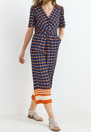 Femme portant une robe midi à motifs bleus, orange et blancs avec manches courtes et sandales plates argentées à lanières, marchant sur un fond blanc.