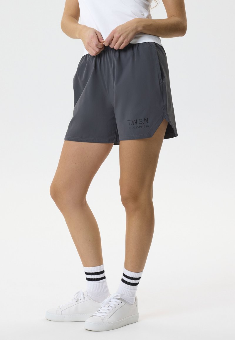 Frau trägt dunkelgraue Sportshorts mit dem Schriftzug T.W.S.N., weiße Turnschuhe und weiße Socken mit schwarzen Streifen, sie justiert den Hosenbund.