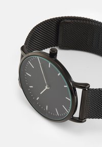 Montre noire avec un bracelet en maille, boîtier rond, design minimaliste, marqueurs d'heure argentés, et un rebord bleu contrastant autour du verre.