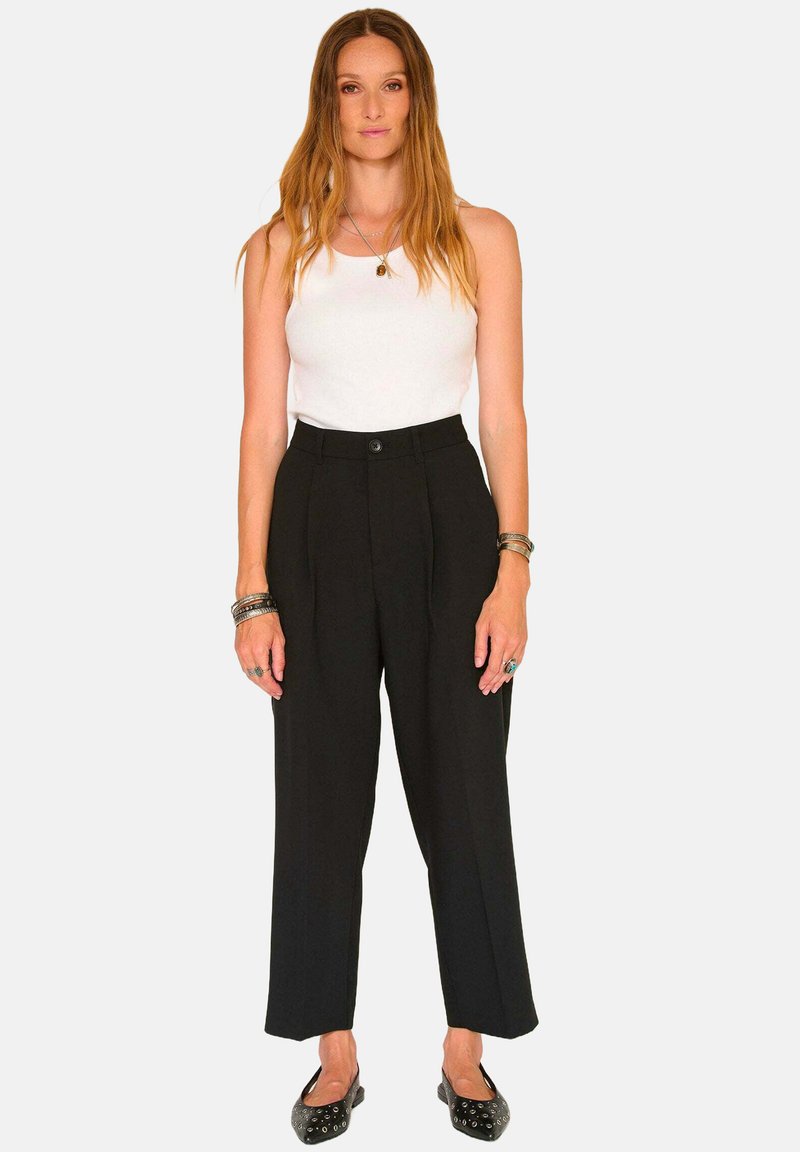 Pantalons noirs taille haute avec un design ajusté, plis à l'avant et fermeture à bouton. Associés à un débardeur blanc et des chaussures noires.