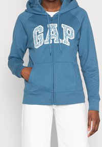 Felpa blu con cerniera e cappuccio regolabile, logo "GAP" bianco. Presenta tasche anteriori e polsini a costine. Tessuto in misto cotone con una consistenza liscia.