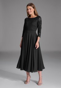 Robe noire avec un corsage en dentelle et des manches trois-quarts, accompagnée d'une jupe plissée fluide. Assortie à des talons à brides argentés.