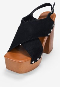 Los tacones de plataforma de ante negro presentan un diseño de tiras cruzadas, suela de madera marrón y acentos plateados en los laterales. Ajustados con una correa de talón.