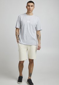 Grau Baumwoll-T-Shirt mit kurzen Ärmeln und Logo auf der Brust, kombiniert mit elfenbeinfarbenen Shorts und schwarzen Sneakers; Tattoos an den Armen sichtbar.