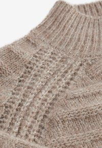 Jasnobrązowy sweter dzianinowy z prążkowanym dekoltem. Posiada teksturowe wzory oraz miękką, puszystą mieszankę wełny.