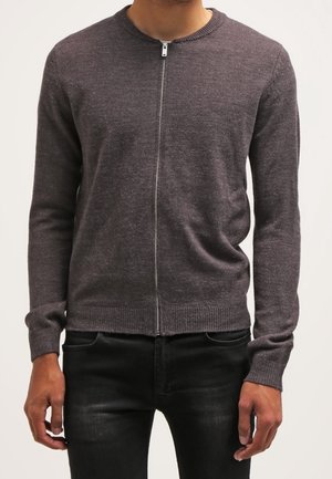 Man wearing a fitted dark gray zip-up sweater and black jeans, standing against a plain light background.