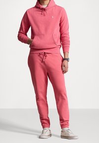 Felpa rosa in cotone con tasca frontale e cappuccio con coulisse, abbinata a pantaloni jogger coordinati con vita elastica e polsini.