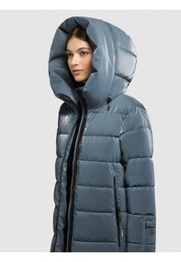 Moteris vilki blizgantį didelio dydžio mėlyną pūstinę striukę su gobtuvu ir juodais užtrauktuko detalėmis ant šviesiai pilkos fono.