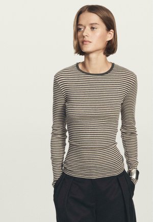 Donna con capelli corti castani che indossa una camicia a maniche lunghe beige e nera a righe, infilata in pantaloni neri, con le mani nelle tasche, che guarda a sinistra.