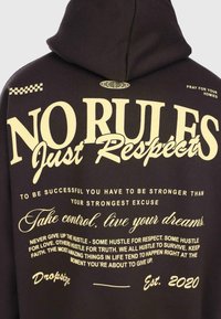 Manteau à capuche noir au dos avec un texte jaune en gras : "PAS DE RÈGLES Juste du Respect" et des citations motivantes sur le travail acharné, le succès et les rêves, daté Est. 2020.