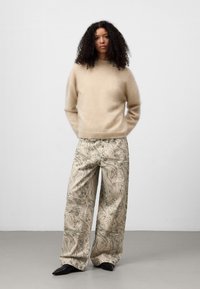 Kobieta z kręconymi włosami w beżowym swetrze, spodniami w szerokie nogawki z wzorem i czarnymi, spiczastymi butami, stojąca na tle białego tła.