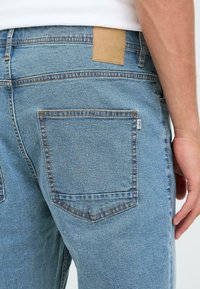 Jeans de mezclilla azul claro con un bolsillo trasero, costuras visibles y una etiqueta de cuero color marrón claro en la cintura. Tejido texturizado con un corte clásico.