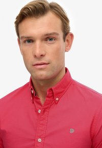 Czerwona koszula z guzikami, z kołnierzykiem, z małym zielonym logo na lewej piersi. Gładka tkanina i regularny krój z widocznymi detalami guzików.