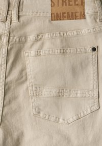 Béžové bavlněné kalhoty se záplatem s nápisem "STREET ONEMEN", jednou zadní kapsou se šitými okraji a detaily z kovových nýtů.