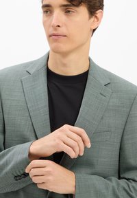 Hellgrüner strukturierten Blazer mit Revers, einer einzelnen Brusttasche und Knöpfen an den Ärmeln, getragen über einem schwarzen Hemd.