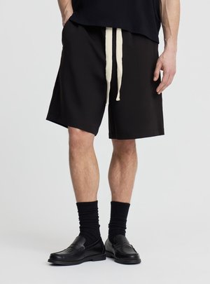 Uomo che indossa pantaloncini neri al ginocchio con cordini bianchi, calzini neri e mocassini in pelle nera, in piedi davanti a uno sfondo semplice.