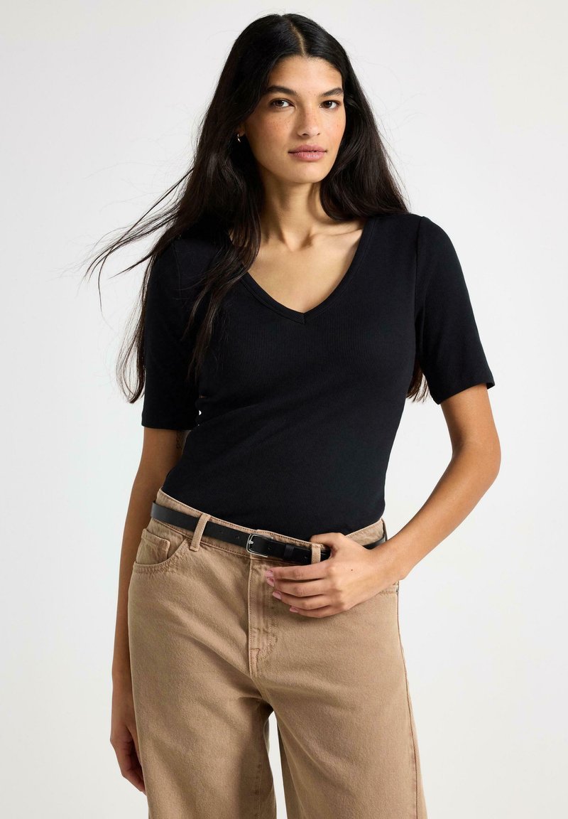 Musta ribbineulottu lyhythihainen V-kauluspaita, yhdistetty vaaleanruskeisiin korkeavyötäröisiin housuihin ja mustaan vyöhön, rento istuvuus.