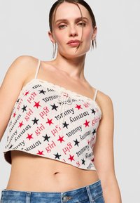 Kremfarget strikket crop-top med spredte svarte og røde stjerner og tekst. Har spaghettitrær og en scalloped kant, med en knyte-detalj foran.