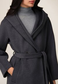 Cappotto grigio texturizzato con colletto a scialle, bottoni doppi e vita con cintura, indossato sopra un dolcevita grigio chiaro.