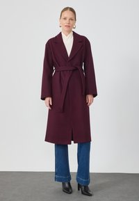 Bordeaux lang frakk med brede slag, belte i midjen og sidelommer. Brukt over en hvit topp og kombinert med vide blå jeans og svarte hæler.