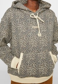 Sudadera con capucha de estampado de leopardo con cordón beige, bolsillo tipo canguro y puños acanalados. La tela tiene una textura suave y un ajuste relajado.